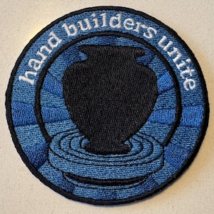May include: A blue and black embroidered patch with the text "Hand Builders Unite" and a black silhouette of a vase.