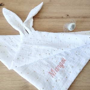 Peut inclure: Une couverture de sécurité pour bébé en mousseline blanche avec des pois dorés, deux longues oreilles de lapin nouées et le prénom Margot brodé en fil rose, accompagnée d'une sucette en silicone transparent avec un motif animal gris.