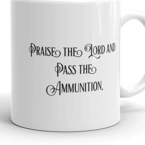 Pode incluir: Caneca de cerâmica branca com uma alça curva. A caneca apresenta o texto "Praise the Lord and Pass the Ammunition" em preto, com uma escrita estilizada. A caneca é projetada para bebidas quentes.