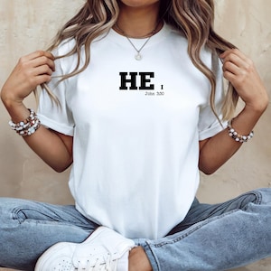 Puede incluir: Camiseta blanca con la palabra "HE" en letras negras en negrita, seguida de una coma, y el texto "John 3:30" en una fuente más pequeña. La camiseta se usa con vaqueros azules y zapatillas blancas.