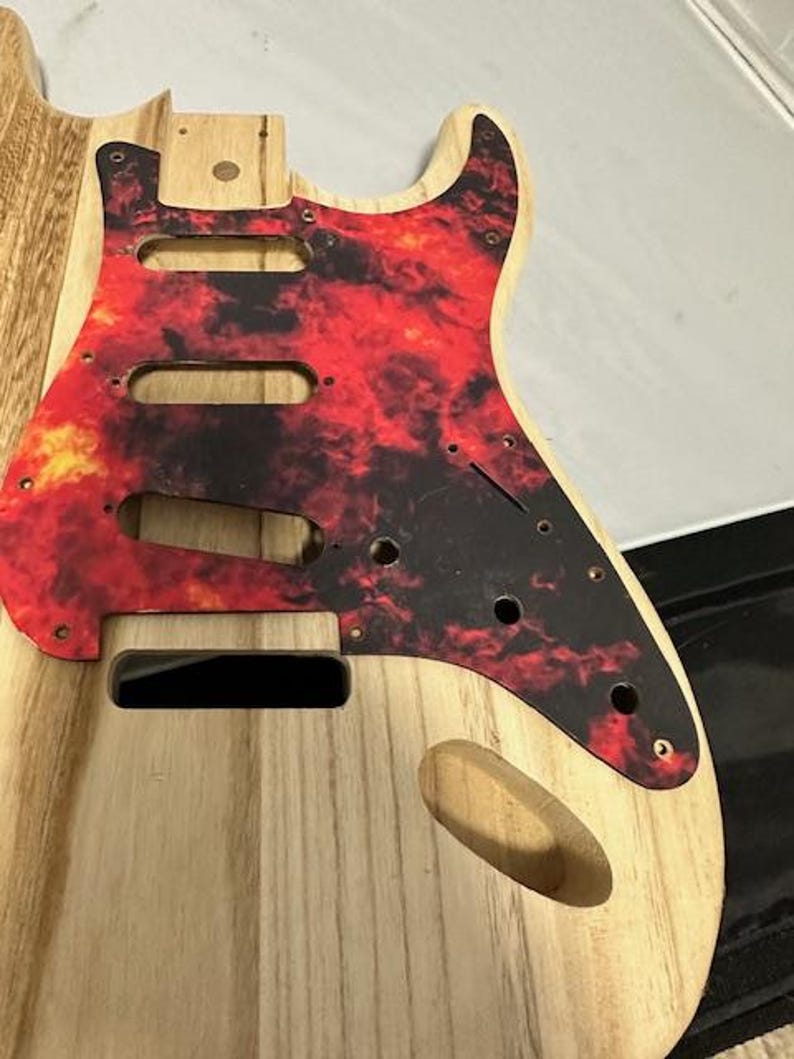 May include: Unfinished wooden guitar body with a red and black flame pattern pickguard.