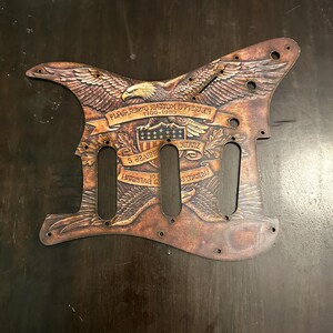 May include: Brown guitar pickguard with an engraved eagle design. The pickguard features the American flag, text, and the years 1900-1953. The pickguard has holes for the guitar's pickups and screws.
