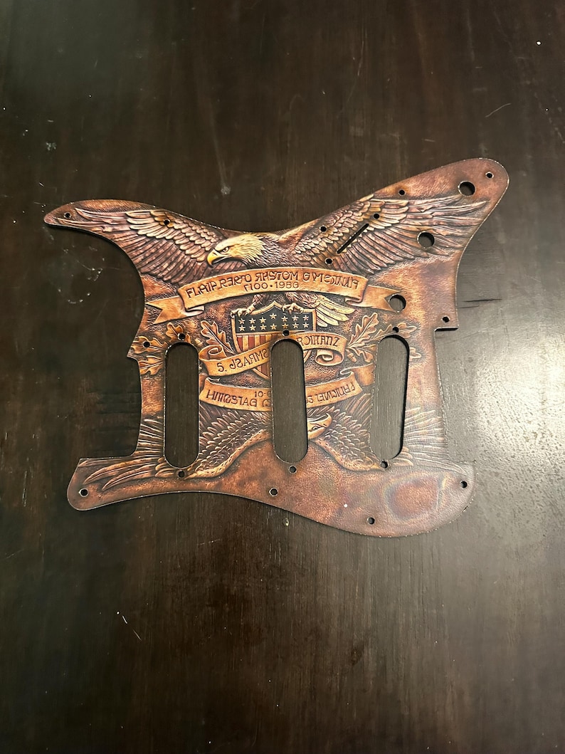May include: Brown guitar pickguard with an engraved eagle design. The pickguard features the American flag, text, and the years 1900-1953. The pickguard has holes for the guitar's pickups and screws.