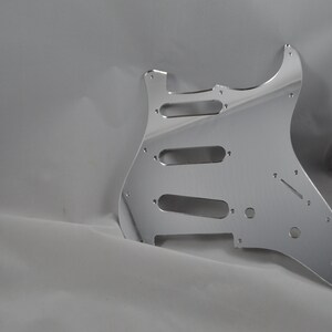 Op de afbeelding: Een zilveren gitaar pickguard met uitsparingen voor pickups en bedieningselementen.