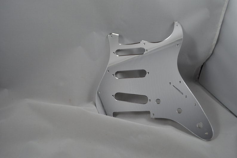Op de afbeelding: Een zilveren gitaar pickguard met uitsparingen voor pickups en bedieningselementen.