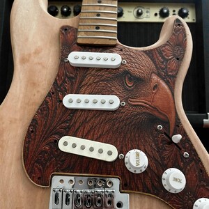 May include: A custom electric guitar with a natural wood finish and a pickguard featuring a detailed eagle design in shades of brown. The guitar has white pickups, control knobs, and a silver bridge. The headstock and fretboard are visible.
