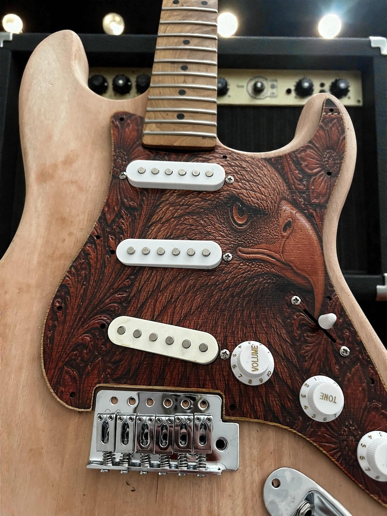 May include: A custom electric guitar with a natural wood finish and a pickguard featuring a detailed eagle design in shades of brown. The guitar has white pickups, control knobs, and a silver bridge. The headstock and fretboard are visible.