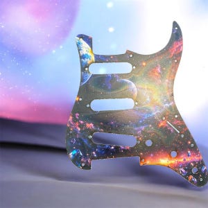 May include: A guitar pickguard with a vibrant galaxy design. The pickguard features a cosmic scene with planets, stars, and nebulae in shades of blue, orange, and purple. It has three pickup cutouts and several screw holes for mounting.