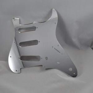 Op de afbeelding: Een chromen gitaar pickguard met uitsparingen voor drie pickups en een regelknop.
