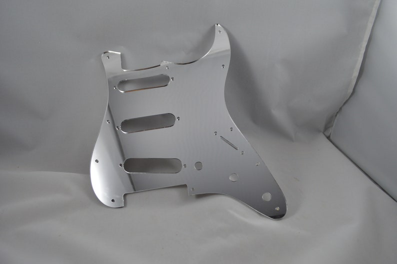 Op de afbeelding: Een chromen gitaar pickguard met uitsparingen voor drie pickups en een regelknop.
