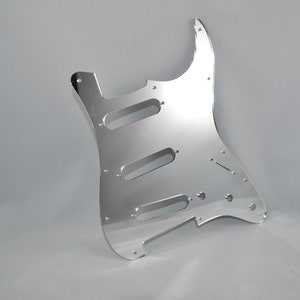 Op de afbeelding: Een zilveren gitaar pickguard met uitsparingen voor pickups en bedieningselementen. De pickguard is gemaakt van een reflecterend materiaal en heeft een gladde, gepolijste afwerking.
