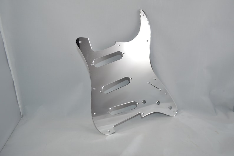 Op de afbeelding: Een zilveren gitaar pickguard met uitsparingen voor pickups en bedieningselementen. De pickguard is gemaakt van een reflecterend materiaal en heeft een gladde, gepolijste afwerking.