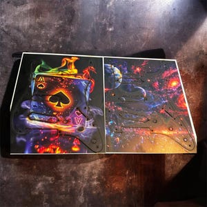 May include: Two guitar pickguards with unique designs. One features the Ace of Spades card engulfed in flames, with vibrant colors. The other depicts a cosmic scene with planets and nebulae. Both have black screw holes.