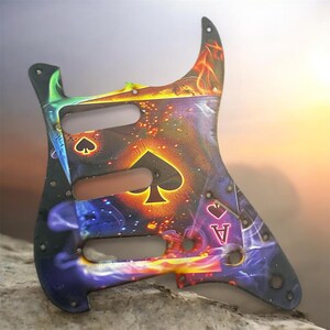 May include: A guitar pickguard with a vibrant design featuring the Ace of Spades card. The pickguard has a black base with a colorful, fiery background and card suit symbols. The design includes the Ace of Spades, Ace of Hearts, and other card suit symbols.