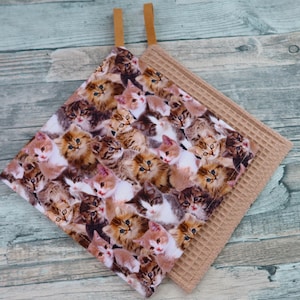 Könnte beinhalten: Zwei quadratische Küchen-Topflappen. Einer zeigt ein sich wiederholendes Muster aus flauschigen Kätzchen in verschiedenen Farben. Der andere ist einfarbiges, beigefarbenes Waffelgewebe. Beide haben Schlaufen aus beigem Stoff zum Aufhängen.