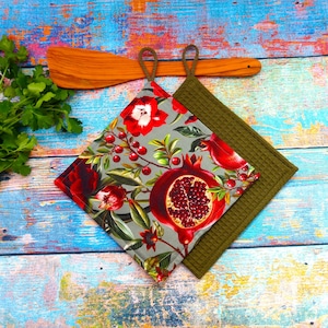 Könnte beinhalten: Zwei quadratische Topflappen. Einer hat einen grauen Hintergrund mit einem floralen Muster aus roten Blumen, Granatäpfeln und grünen Blättern. Der andere ist olivgrün. Beide haben eine Schlaufe zum Aufhängen. Ein Holzspatel liegt darüber.