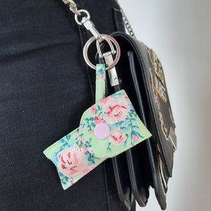 May include: A small, floral-patterned keychain pouch in shades of green, pink, and blue, attached to a silver chain and a black handbag. The pouch has a pink button closure and a loop for the keychain ring. The floral design is a repeating pattern.