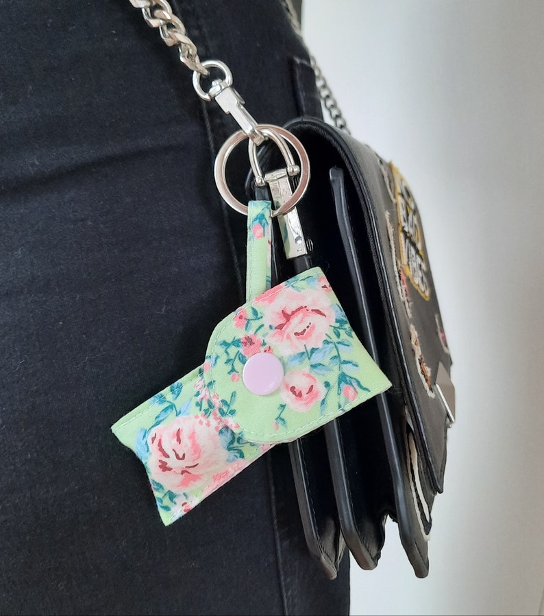 May include: A small, floral-patterned keychain pouch in shades of green, pink, and blue, attached to a silver chain and a black handbag. The pouch has a pink button closure and a loop for the keychain ring. The floral design is a repeating pattern.