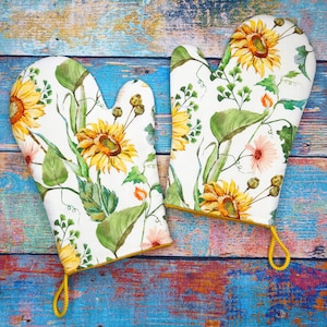 Könnte beinhalten: Ein Paar weiße Ofenhandschuhe mit einem lebendigen Blumenmuster mit Sonnenblumen, grünen Blättern und rosa Blüten. Die Handschuhe haben eine gelbe Paspel und eine gelbe Schlaufe zum Aufhängen. Der Hintergrund ist eine rustikale, mehrfarbige Holzoberfläche.