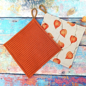 Könnte beinhalten: Zwei quadratische Topflappen. Einer ist einfarbig orange mit Waffelstruktur und einer braunen Schlaufe. Der andere ist beige mit einem Muster aus orangefarbenen Lampionblumen und einer braunen Schlaufe. Die Topflappen liegen auf einer bunten Holzoberfläche.