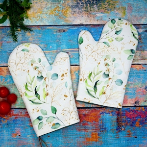 Könnte beinhalten: Ein Paar weiße Ofenhandschuhe mit einem botanischen Design. Die Handschuhe zeigen grüne und blaue Blätter mit goldenen Akzenten. Der Hintergrund ist eine rustikale blau-braune Holzoberfläche.