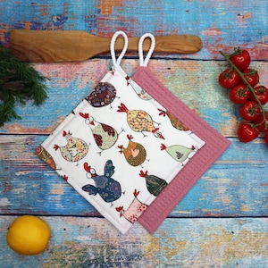 Könnte beinhalten: Zwei quadratische Topflappen. Einer ist weiß mit bunten Hühnerdesigns, der andere ist einfarbig mauve. Beide haben weiße Schlaufengriffe. Die Topflappen liegen auf einer blau- und holzfarbenen Oberfläche, mit einem Kochlöffel, Tomaten und einer Zitrone.