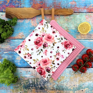 Könnte beinhalten: Zwei Küchen-Topflappen. Einer ist weiß mit einem floralen Rosenmuster in Rosa- und Rottönen. Der andere ist einfarbig rosa mit einer Waffelstruktur. Beide haben braune Lederaufhängeschlaufen.