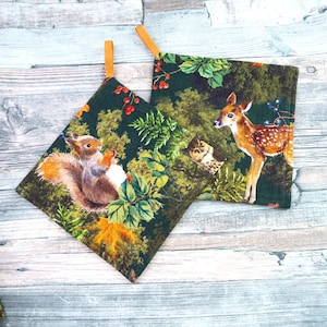Könnte beinhalten: Zwei quadratische Topflappen mit Waldtiermotiven. Einer zeigt ein Eichhörnchen, der andere ein Reh. Beide haben einen dunkelgrünen Hintergrund mit Blättern, Beeren und einer Eule. Jeder hat eine beige Schlaufe zum Aufhängen.