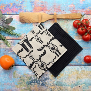 Könnte beinhalten: Zwei quadratische Küchen-Topflappen. Einer ist schwarz, der andere beige mit einem schwarzen Elchmuster. Ein Holzlöffel und ein Zweig Tomaten sind im Hintergrund. Die Topflappen liegen auf einer blau und holzfarbenen Oberfläche.