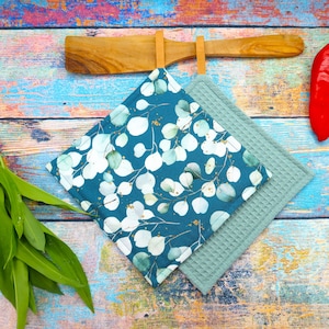 Könnte beinhalten: Zwei quadratische Topflappen. Einer ist türkis mit einem weißen und grünen Eukalyptusblattmuster. Der andere ist einfarbig hellgrün gewebt. Eine hölzerne Löffelablage befindet sich über den Topflappen. Der Hintergrund ist eine farbenfrohe Holzoberfläche.