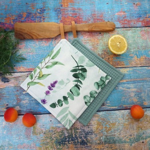 Könnte beinhalten: Zwei quadratische Küchen-Topflappen. Einer ist hellgrün gewebt, der andere ist weiß mit einem Aquarell-Eukalyptus- und Lavendel-Design. Ein Holzspatel hängt darüber. Aprikosen und eine Zitronenhälfte sind ebenfalls im Bild.