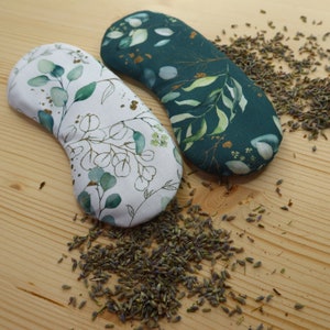 Könnte beinhalten: Zwei Augenpads mit botanischen Mustern liegen auf einer Holzoberfläche, umgeben von getrocknetem Lavendel. Ein Pad hat einen weißen Hintergrund mit grünen Eukalyptusblättern, das andere einen dunkelgrünen Hintergrund mit ähnlichem Laub.