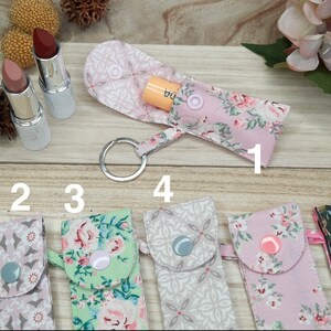 May include: Fabric lipstick holders with keychains, in floral and geometric patterns. The colours are pink, green, and grey. Two lipsticks are visible. The holders are closed with snap buttons. The image is on a wooden surface.