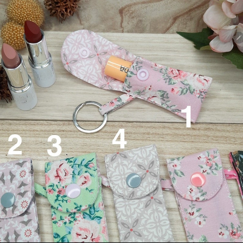 May include: Fabric lipstick holders with keychains, in floral and geometric patterns. The colours are pink, green, and grey. Two lipsticks are visible. The holders are closed with snap buttons. The image is on a wooden surface.