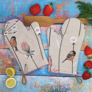 Könnte beinhalten: Ein Paar beige Ofenhandschuhe mit Blumen- und Vogelmuster. Die Handschuhe zeigen einen braunen Vogel, der auf einem Stiel mit rosa und weißen Blüten sitzt. Sie haben eine rosa Einfassung und eine braune Schlaufe zum Aufhängen. Erdbeeren und Zitronen im Hintergrund.