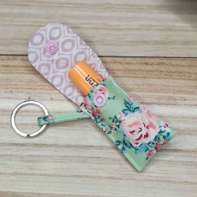 May include: A fabric lip balm holder with a key ring, featuring a floral print. The holder is open, displaying a lip balm tube. The holder's exterior has a light green background with a pink floral design. The interior has a pink and white geometric pattern.