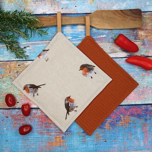 Könnte beinhalten: Zwei quadratische Topflappen. Einer ist beige mit gestickten Rotkehlchen, der andere ist strukturiert orange. Beide haben Lederschlaufen zum Aufhängen. Rote Paprika und Rosmarinzweige liegen auf einer blau-holzmaserierten Oberfläche.