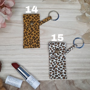 May include: Two lipstick holders with keychains, one in a tan and brown leopard print and the other in a white and brown leopard print. A tube of red lipstick and its cap are also visible.