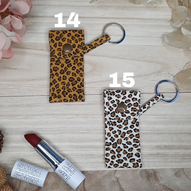 May include: Two lipstick holders with keychains, one in a tan and brown leopard print and the other in a white and brown leopard print. A tube of red lipstick and its cap are also visible.