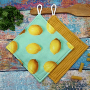 Könnte beinhalten: Zwei quadratische Topflappen. Einer ist helltürkis mit einem Muster aus gelben Zitronen. Der andere ist senfgelb mit Waffelwebung. Beide haben weiße Aufhängeschlaufen. Der Hintergrund ist eine rustikale, mehrfarbige Holzoberfläche.