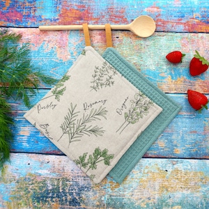 Könnte beinhalten: Zwei quadratische Topflappen. Einer ist hellbeige mit grünen Kräuterillustrationen und den Wörtern "Petersilie", "Rosmarin" und "Oregano". Der andere ist türkisblau mit Waffelstruktur. Ein Holzlöffel und Erdbeeren sind ebenfalls zu sehen.