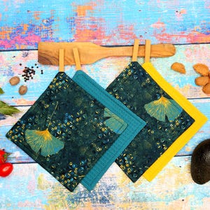 Könnte beinhalten: Zwei quadratische Topflappen mit einem Ginkgo-Blattmuster. Einer ist türkis mit einem türkisfarbenen Rand, der andere ist dunkelgrün mit einem gelben Rand. Sie hängen an einem Holzutensil auf einer farbenfrohen Holzoberfläche.