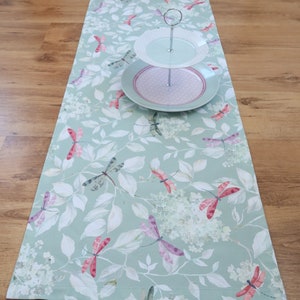 Könnte beinhalten: Hellgrüner Tischläufer mit einem Libellen- und Blumenmuster. Der Läufer ist mit rosa und lila Libellen und weißen Blumen verziert. Ein zweistöckiger Servierständer mit weißen und rosa Tellern steht darauf.