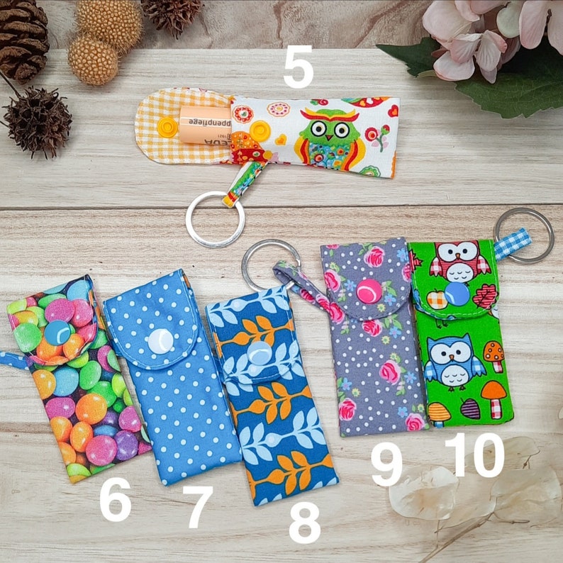 May include: A selection of fabric keychain holders with diverse patterns. Designs feature polka dots, floral motifs, and owl illustrations. Each holder has a snap closure and a metal keyring. Some have text.