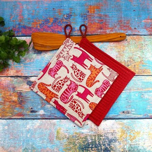 Könnte beinhalten: Zwei quadratische Topflappen. Einer ist rot, der andere beige mit einem sich wiederholenden Muster aus rosa und orangefarbenen Katzen. Ein Holzspatel mit Schlaufengriff befindet sich hinter den Topflappen.