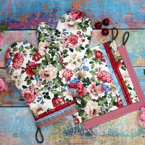 Könnte beinhalten: Ein Set aus Ofenhandschuhen und Topflappen mit Blumenmuster. Das Set hat einen weißen Hintergrund mit roten, rosa und grünen Blumenmustern. Der Topflappen hat eine rote und blaue Spitzenborte und eine braune Schlaufe zum Aufhängen.