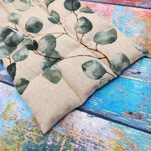 Könnte beinhalten: Ein beiges rechteckiges Wärmekissen mit einem botanischen Eukalyptusblatt-Design. Die Blätter sind ein gedämpftes Grün mit braunen Stielen, gedruckt auf einem leinenartigen Stoff. Das Wärmekissen ist in Abschnitte unterteilt.