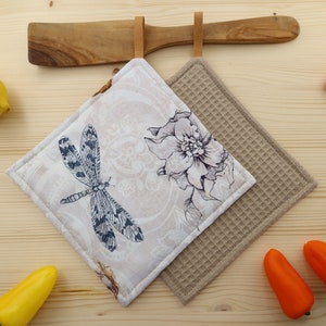 Könnte beinhalten: Zwei quadratische Topflappen. Einer ist beige mit einem Libellen-Design in Marineblau und Weiß. Der andere ist ein strukturierter, hellbrauner Waffelstrick. Ein Kochlöffel und ein Holzhalter befinden sich über den Topflappen. Gelbe und orangefarbene Paprika sind im Hintergrund.