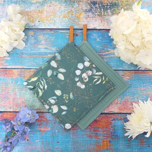 Könnte beinhalten: Zwei quadratische Topflappen. Einer ist türkis mit einem weißen und goldenen Blumenmuster, der andere ist einfarbig türkis. Beide haben braune Lederaufhängeschlaufen. Die Topflappen liegen auf einer blau und holzfarbenen Oberfläche.