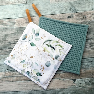 Könnte beinhalten: Zwei quadratische Topflappen. Einer ist weiß mit einem botanischen Design aus grünen Blättern und goldenen Akzenten. Der andere ist türkis mit einer Waffelstruktur. Beide haben braune Lederschlaufen zum Aufhängen.